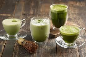Vier Gläser mit Matcha Latte und Tee aus Bio Matchapulver auf Holztisch, Matchabesen daneben